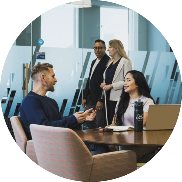 A man and woman are working together at a conference table, talking together. Another man and woman are walking outside the glass-walled room, and another man works at a separate table, his back to the others.