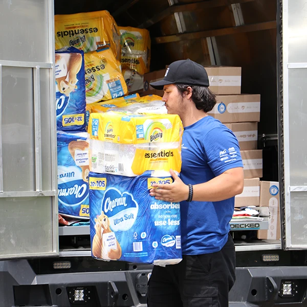 A young man in a black cap holds a pack of Charmin and Bounty while walking in front of an open truck filled with Charmin, Bounty, and P&G disaster relief kits.