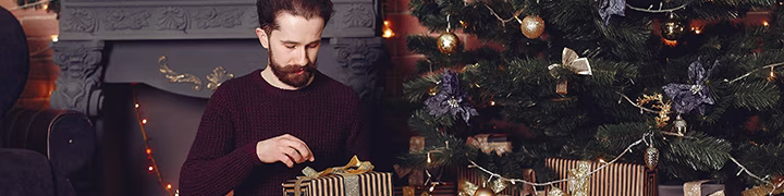 Mann öffnet sein Weihnachtsgeschenk im festlich geschmückten Wohnzimmer | Weihnachtsgeschenke online bestellen shöpping.at