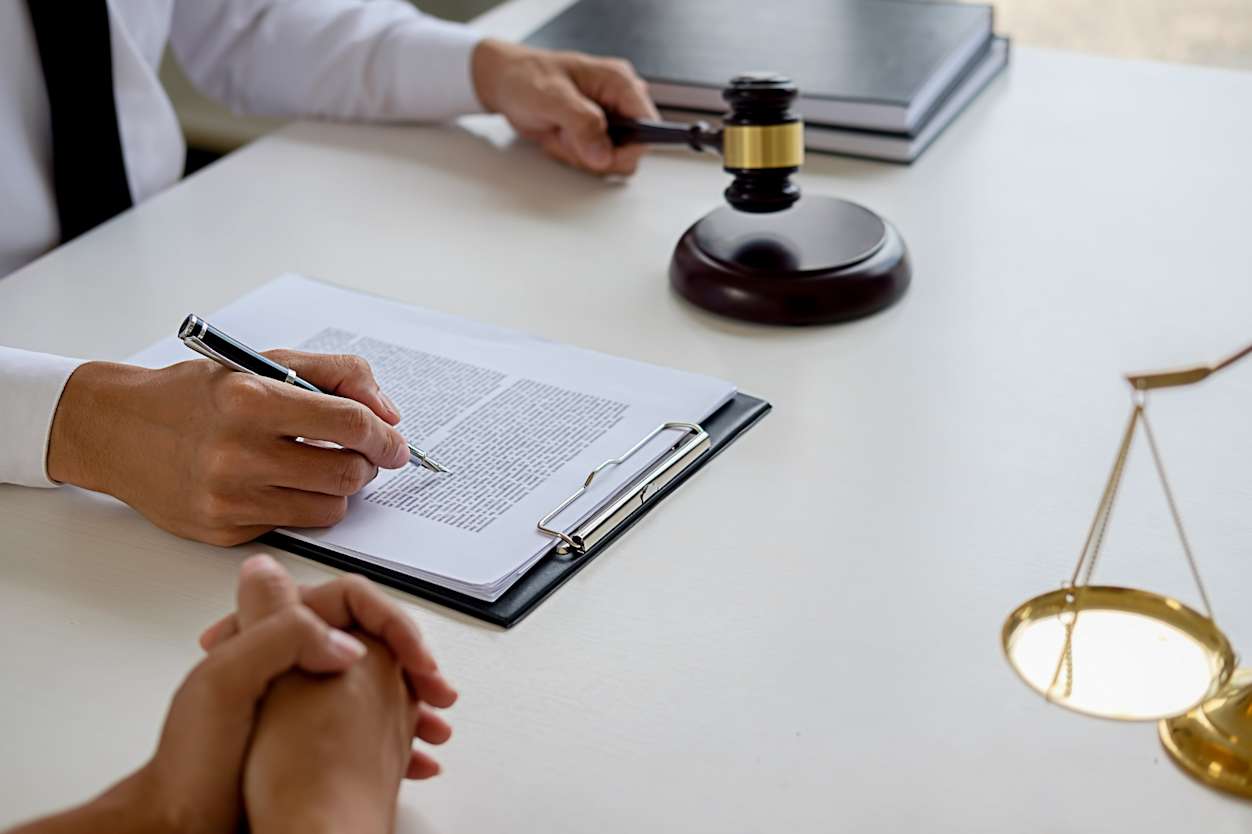 Close up of a person signing a contract holding a gavel, next to a pair of scales.