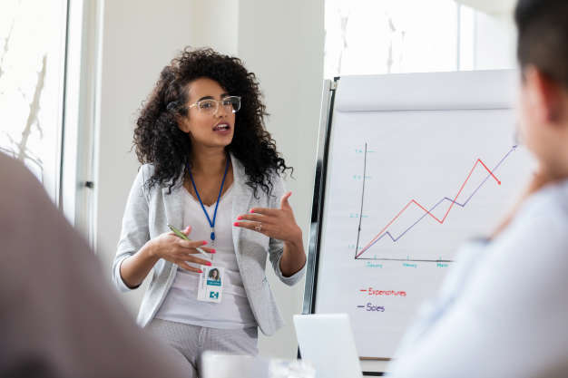 A serious young businesswoman stands before a group of medical professional during a meeting. There is a graph beside her as she explains financial data.