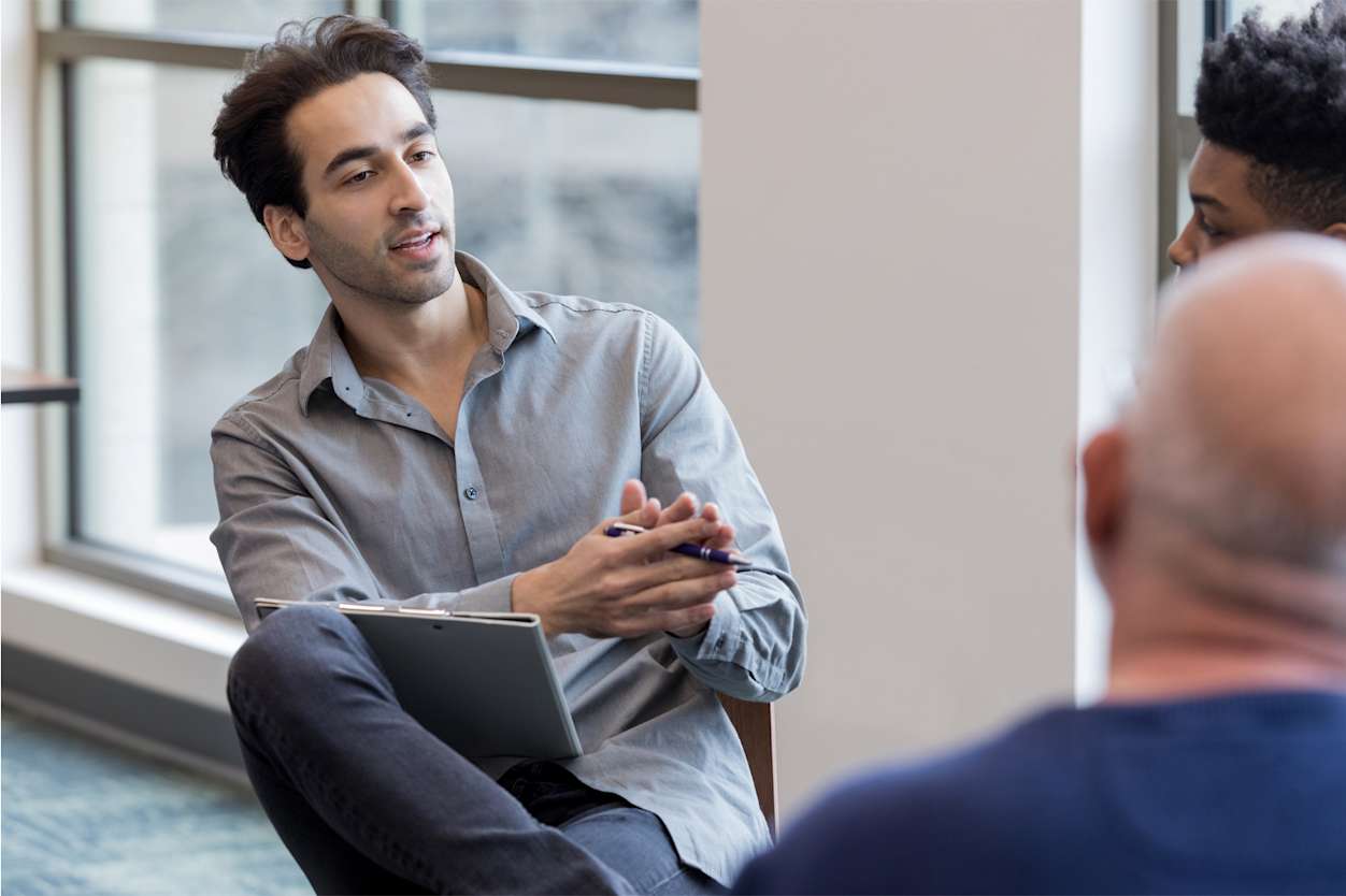 Man sits having a discussion with two other men, he is holding a notebook and pen
