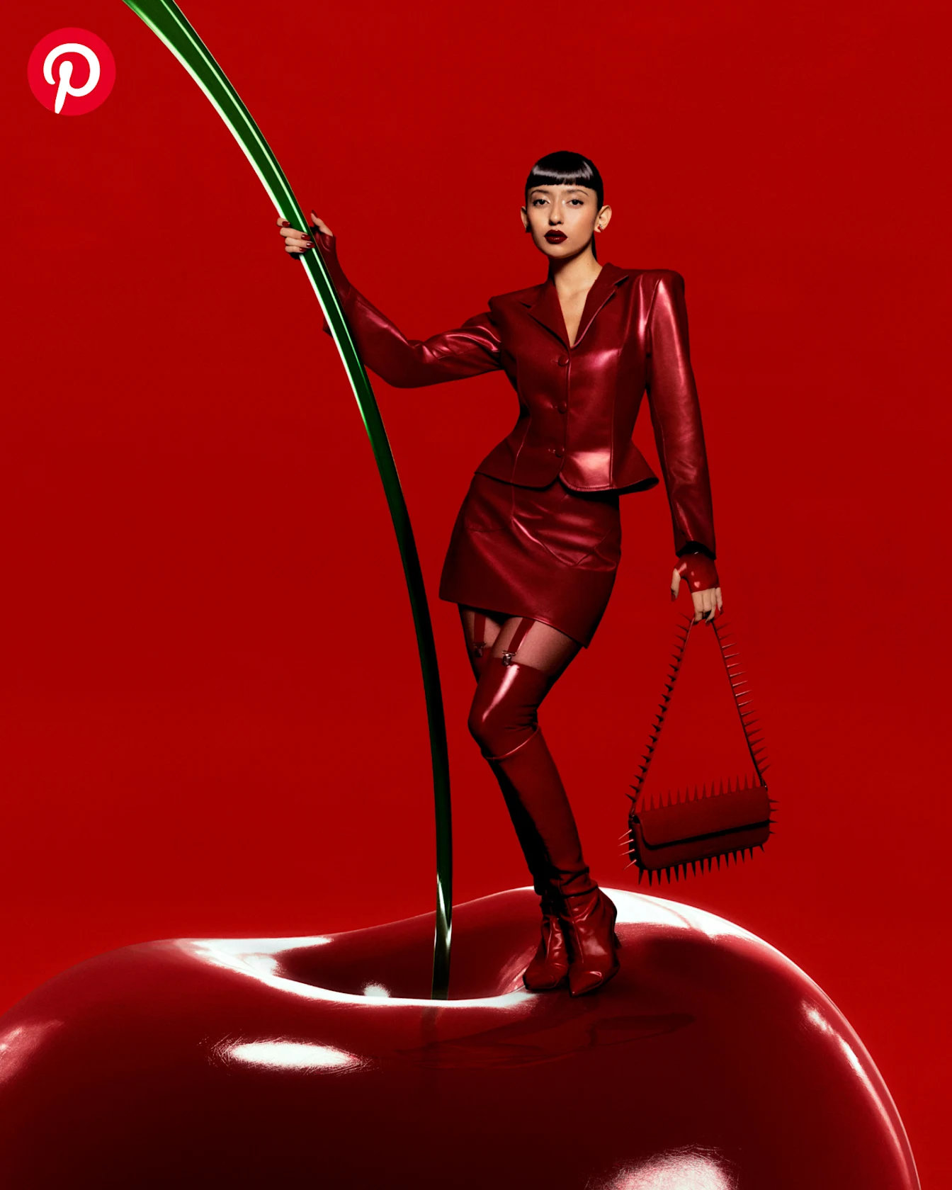 A woman with sleek black hair, dark red lipstick and a head-to-toe red leather outfit poses on top of a giant cherry