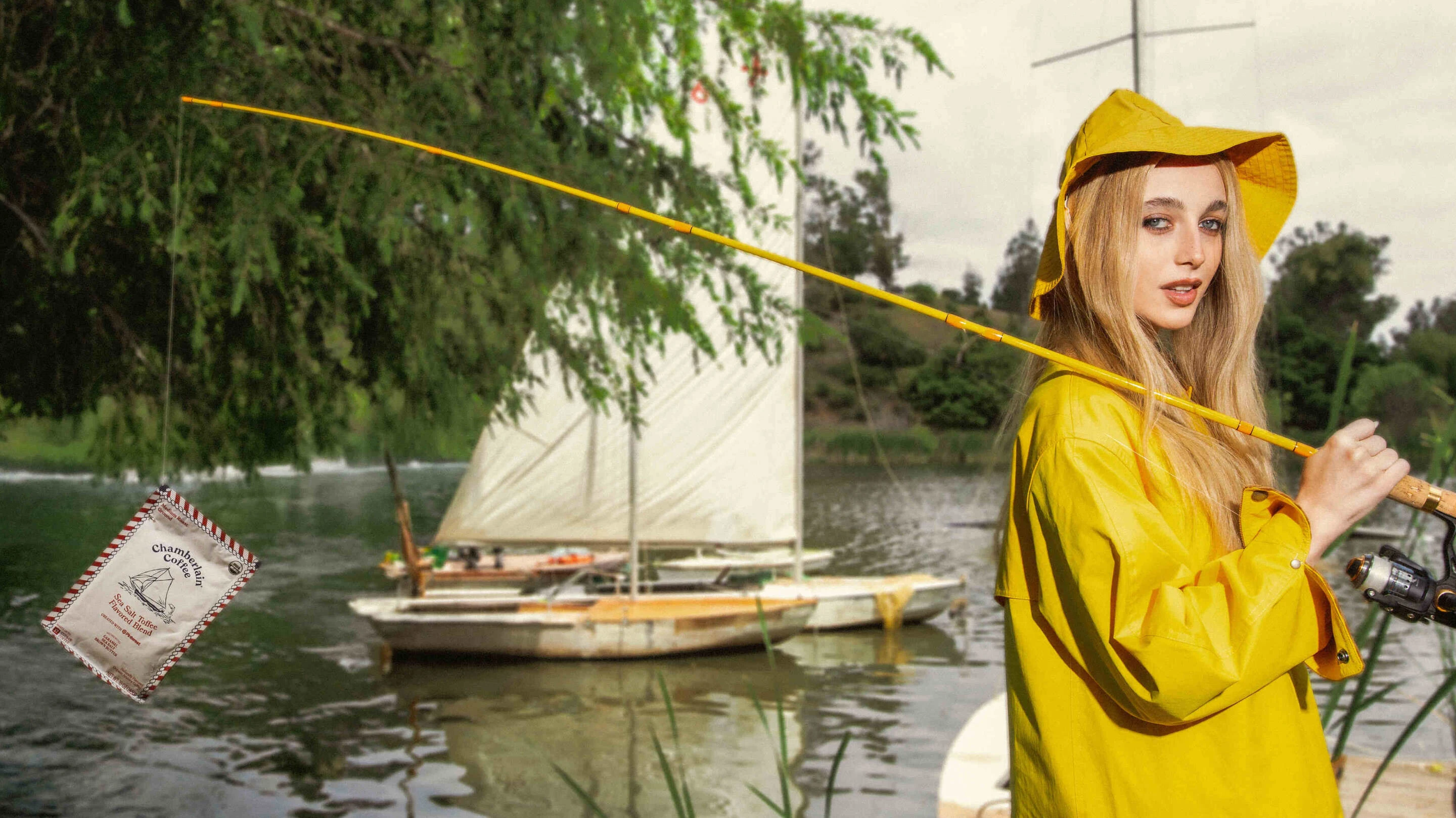 Gen Z creator Emma Chamberlain wears a yellow raincoat and rain hat. A fishing rod is slung over her shoulder with a bag of Chamberlain Coffee’s Sea Salt Toffee flavored coffee blend on the hook.