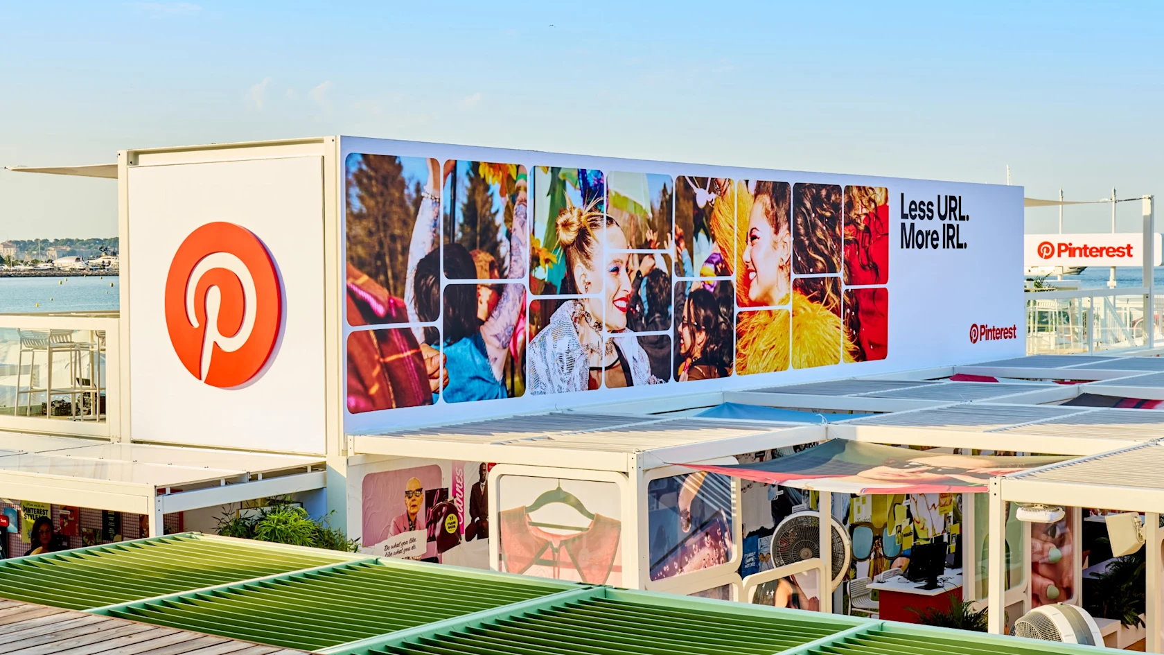 Vista exterior del espacio Manifestival de Pinterest en Cannes, con un gran logotipo de Pinterest y un panel con fotos coloridas y el lema “Less URL. More IRL.” frente a un entorno junto al mar.