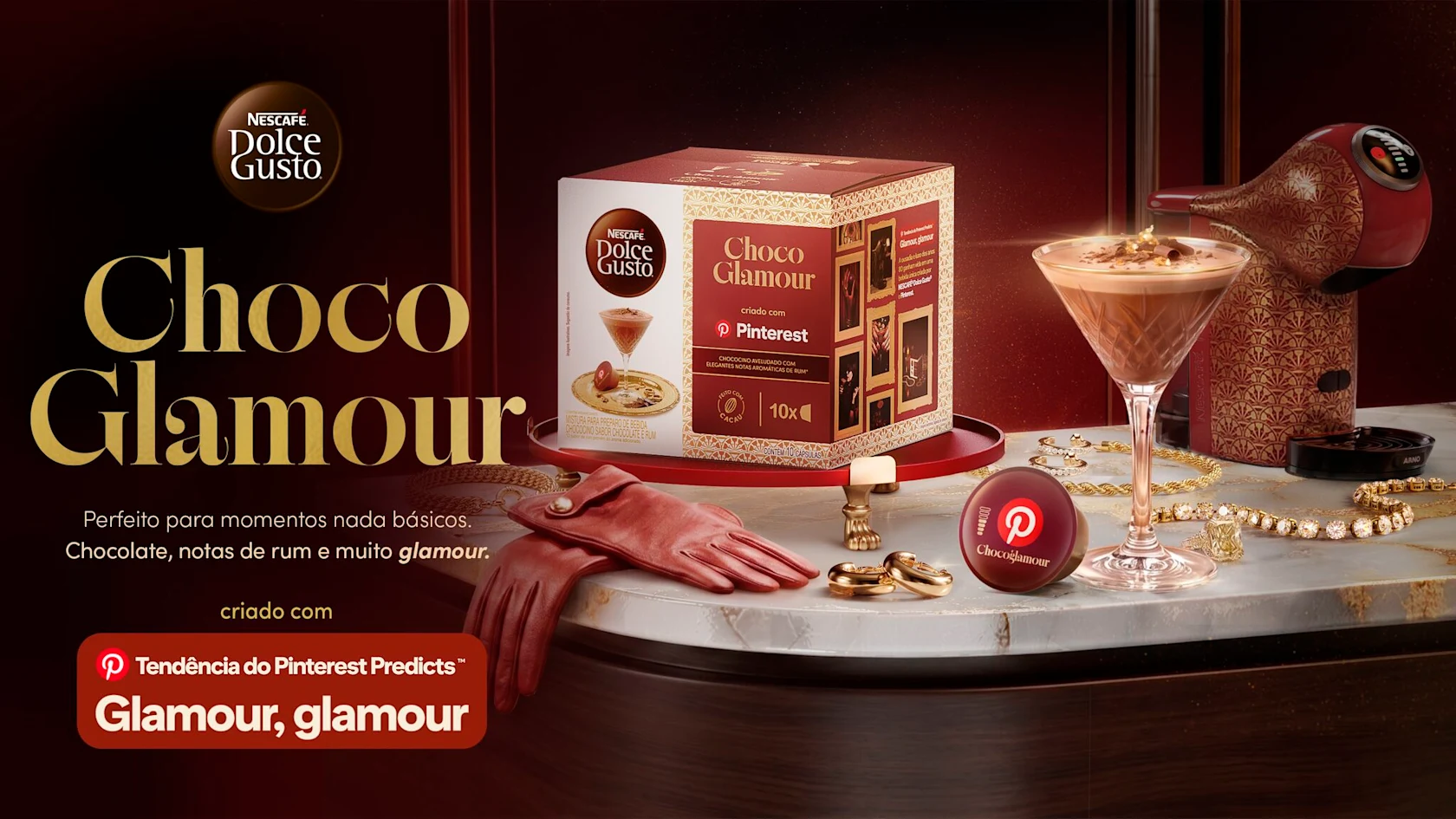 Caixa e máquina NESCAFÉ® Dolce Gusto® Choco Glamour com bebida de chocolate, joias, anéis e luvas vermelhas sobre mesa de mármore.