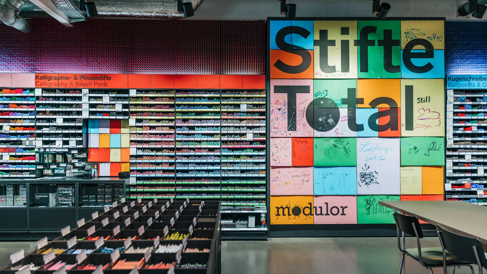 Innenansicht des Modulor-Ladens mit Regalen voller Stifte, Marker und Kunstbedarf. Eine große, bunte Wandinstallation zeigt „Stifte Total" mit dem Modulor-Logo.