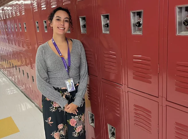 Teacher smiling while standing in a hallway and leaning against a row of red school lockers.