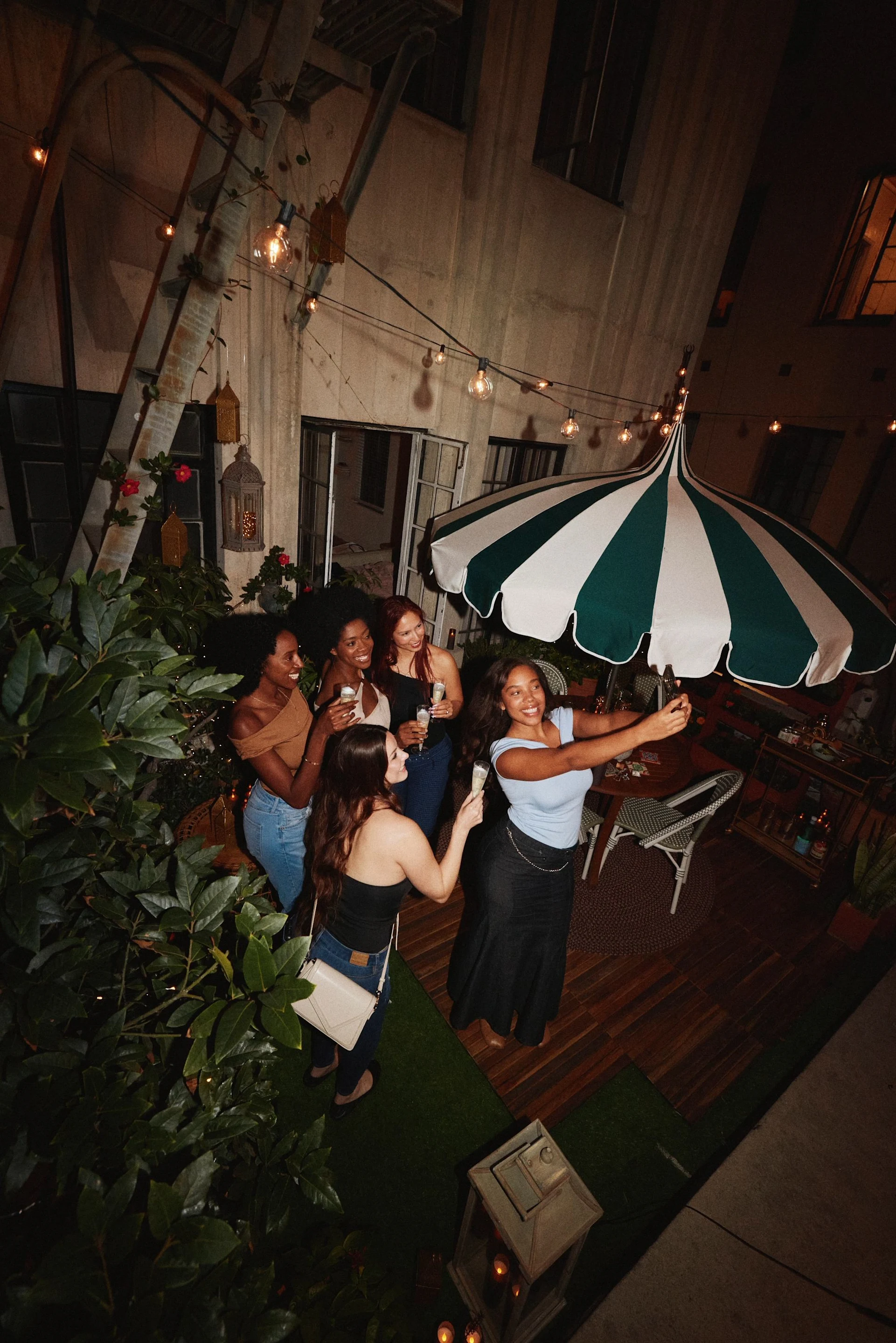 Five friends on a cozy outdoor patio at night with one woman taking a group selfie under string lights and a green-and-white striped umbrella.