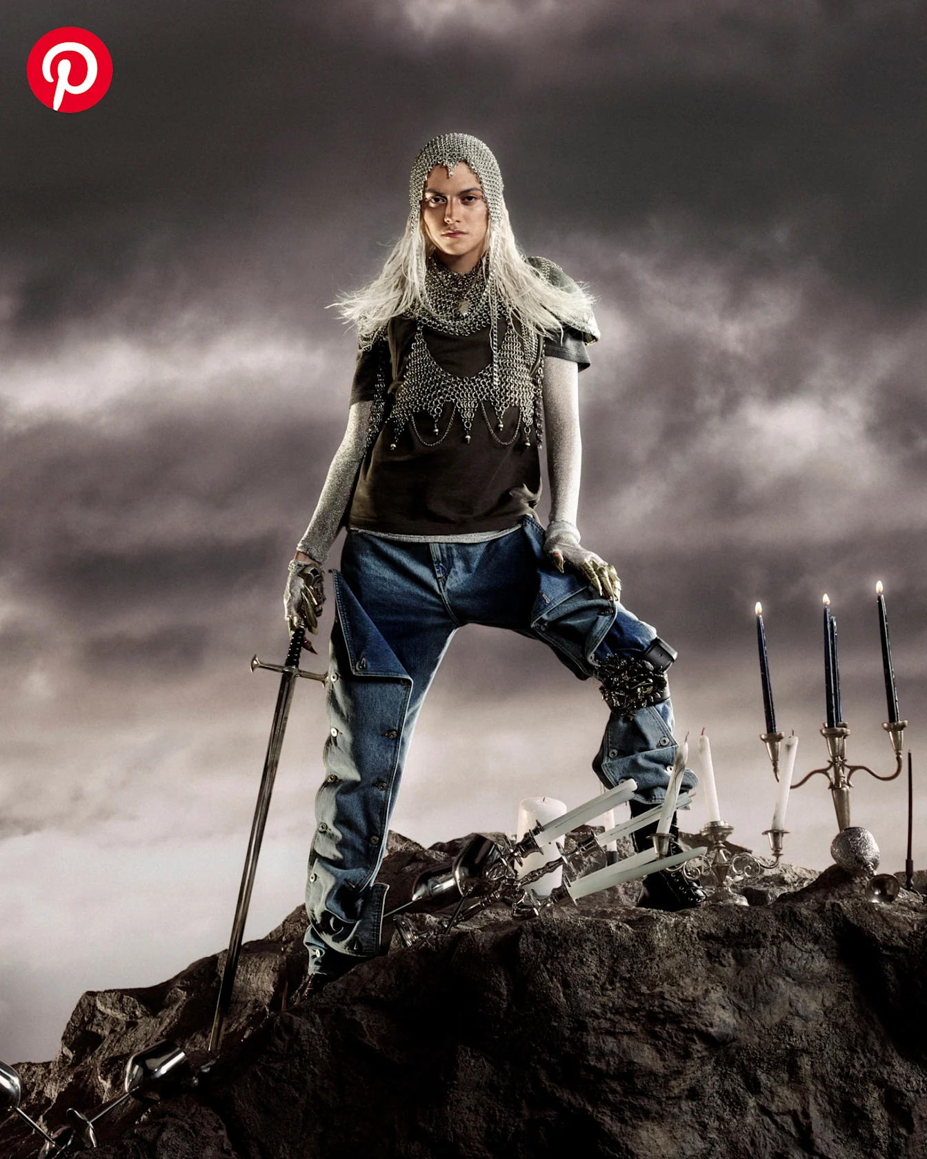 A man wearing a chainmail headpiece and holding a sword poses against a stormy sky surrounded by ornate candelabras