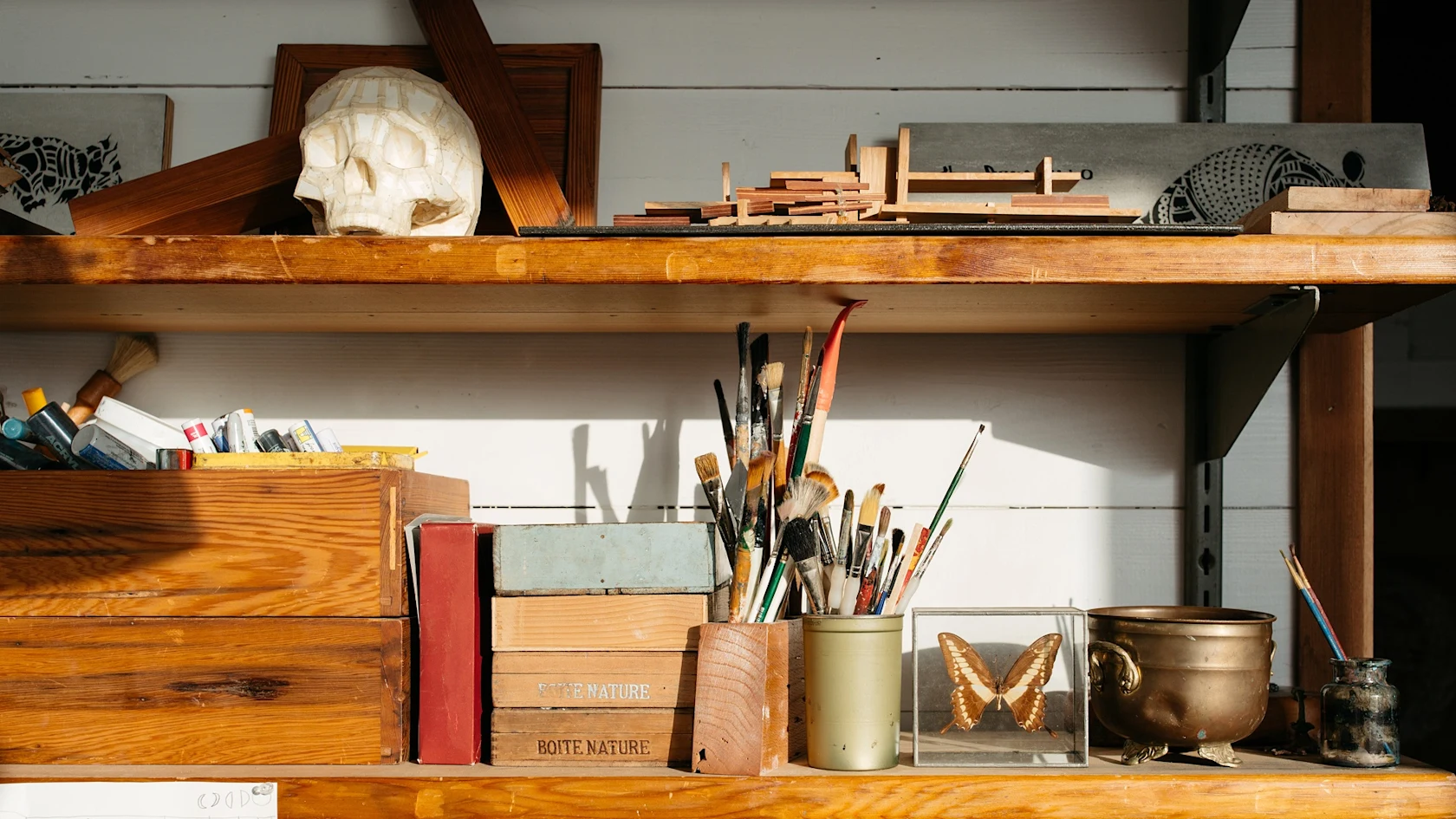 Étagères en bois d’atelier avec pinceaux, boîtes nature, tubes de peinture, crâne sculpté, morceaux de bois, papillon sous verre et bol en laiton.