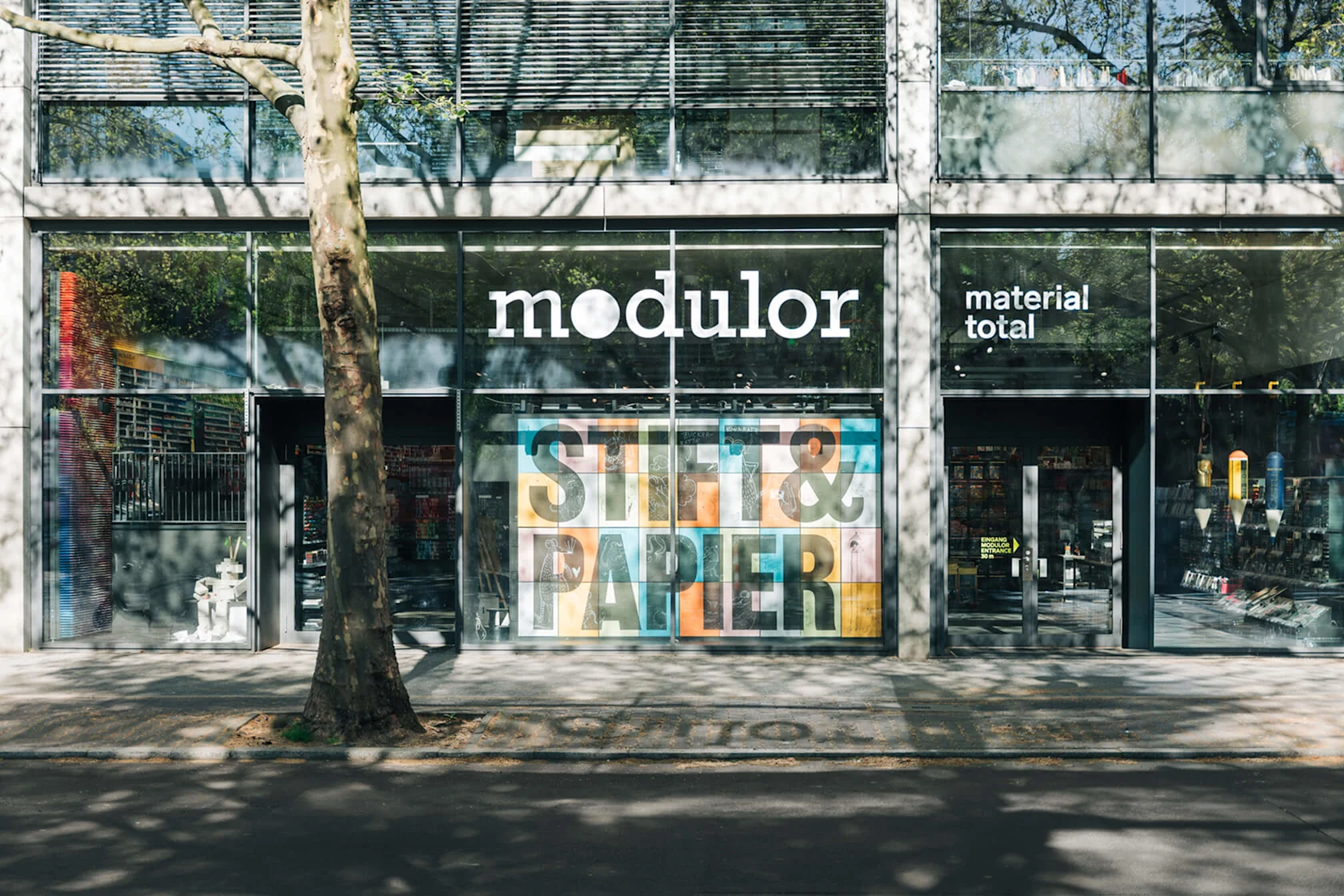 Außenansicht des Modulor-Ladens in Berlin. Die große Glasfassade zeigt die „Stift & Papier"-Kampagne auf bunten Panels.
