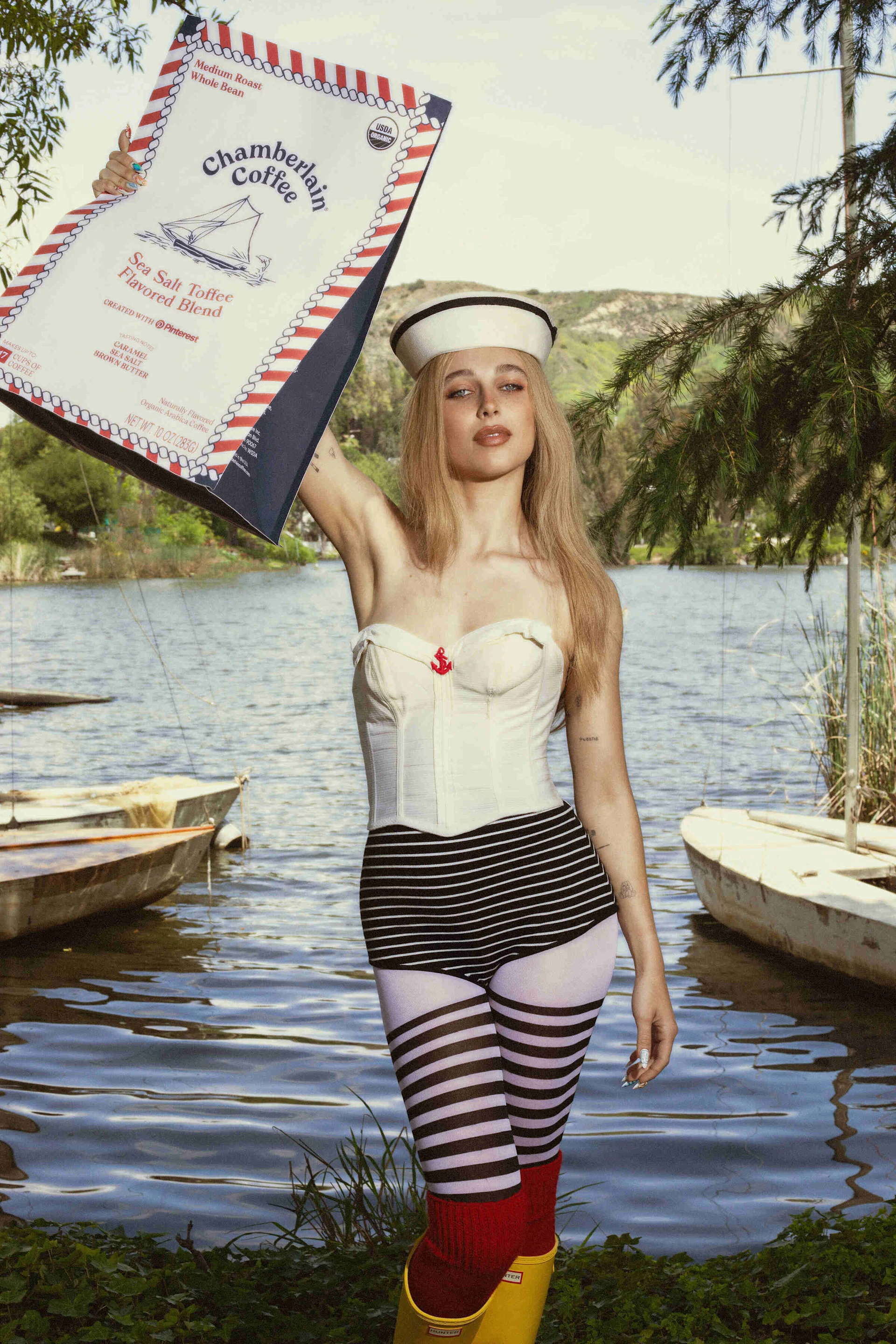 Emma Chamberlain poses in front of a dock wearing a sailor costume and holding up a giant bag of Chamberlain Coffee’s Sea Salt Toffee flavored coffee blend.