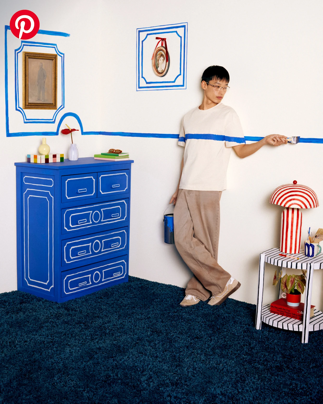 A man stands against a white wall in a white shirt and baggy beige pants. He holds a paintbrush, and there’s a blue stripe across his shirt and the wall. The room contains a blue dresser, a red-striped lamp and a blue-striped side table.