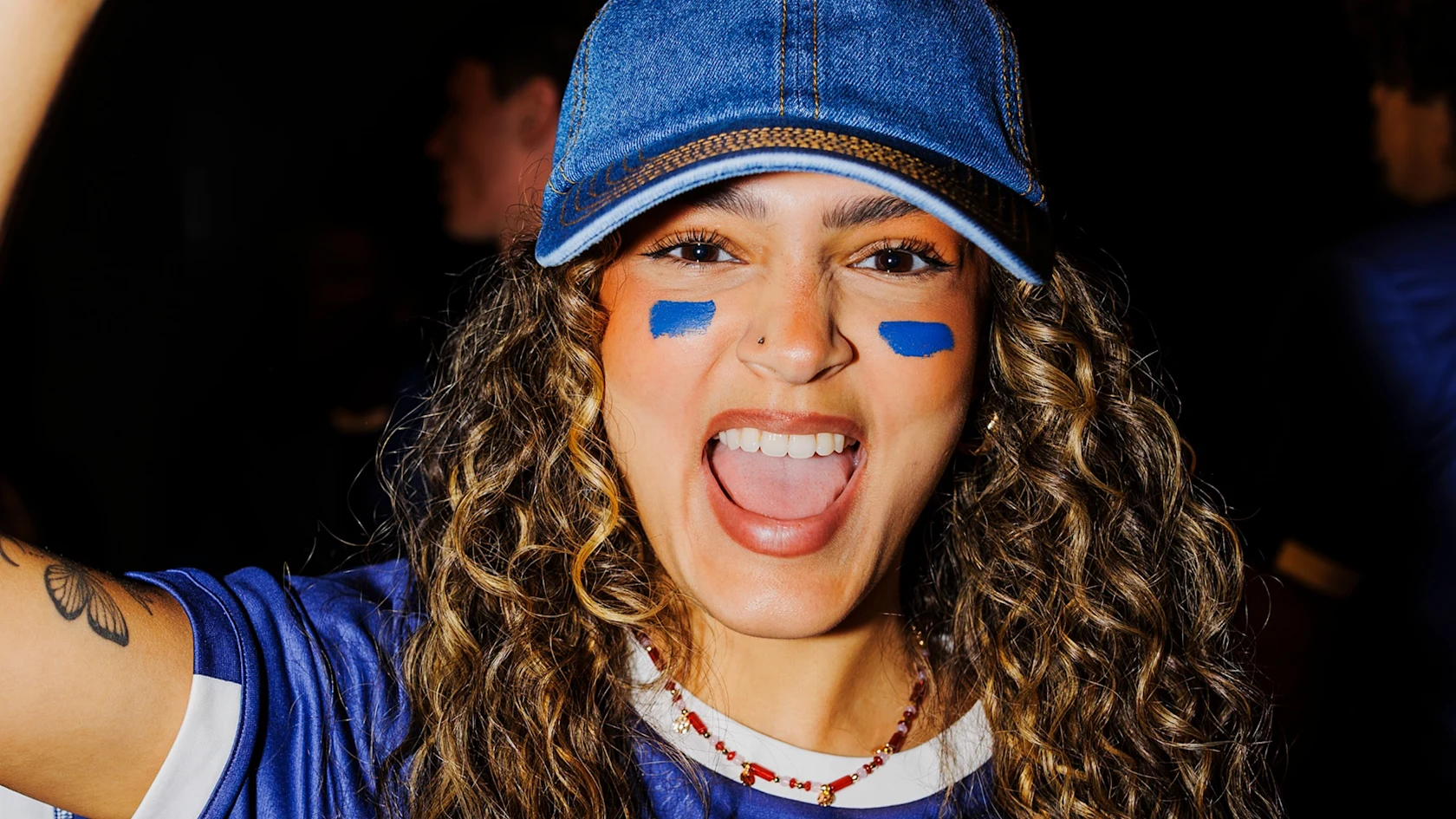 A young woman with curly hair, blue face paint stripes, and a denim cap smiles and cheers energetically, showing team spirit in a sporty blue jersey.