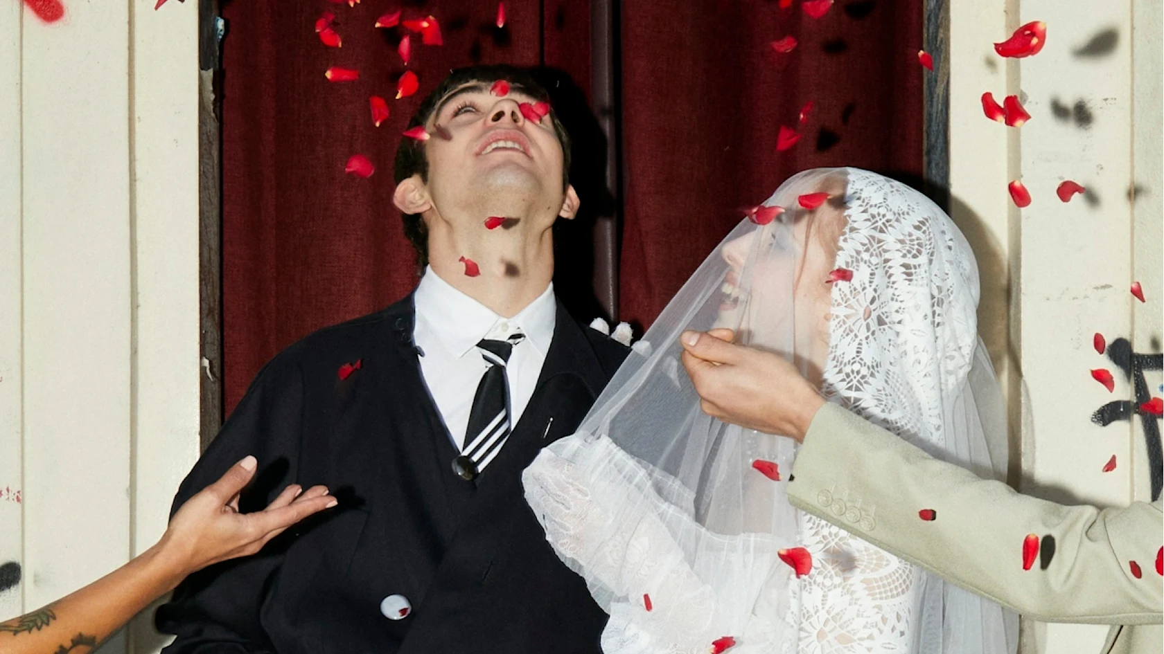 Pareja sonriendo bajo una lluvia de confeti de pétalos rojos; la novia lleva velo y guantes de encaje