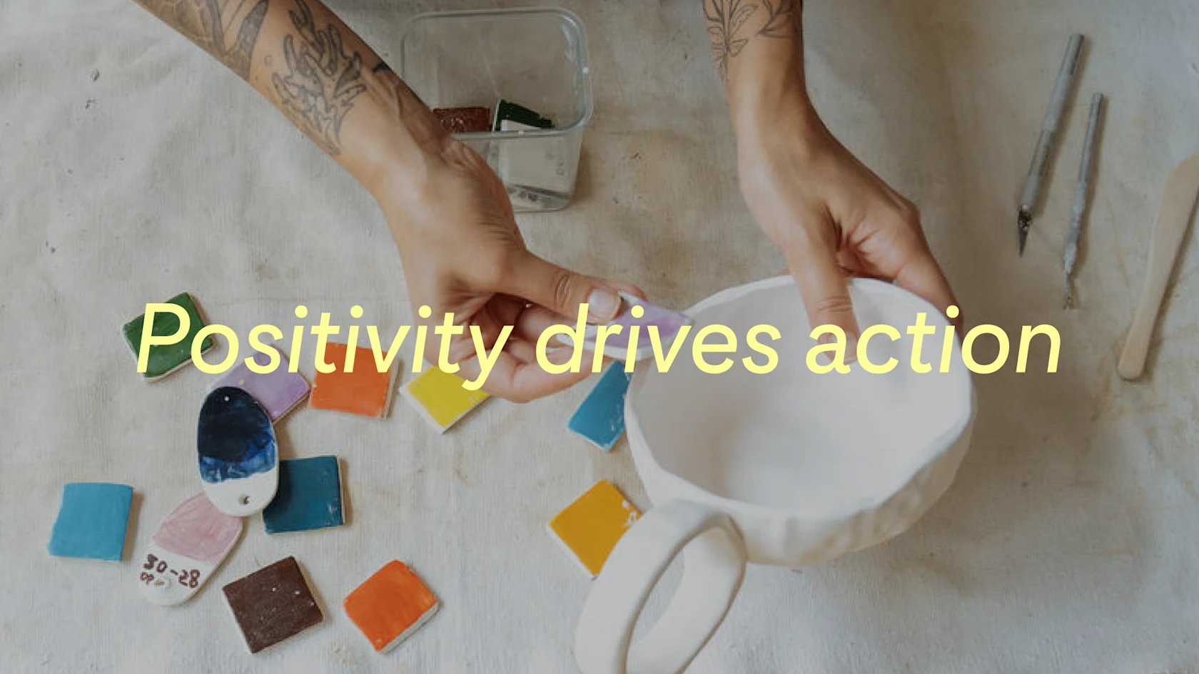 A ceramicist tests different color swatches on a handmade mug, with the words "Positivity drives action" displayed over the image