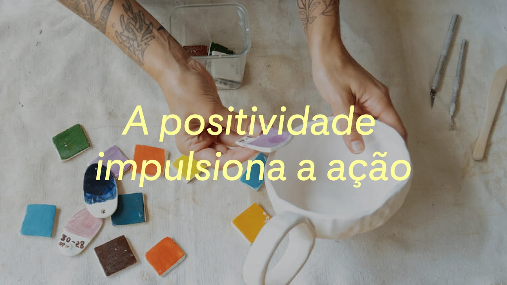 Um ceramista testa diferentes amostras de cor em uma caneca artesanal, com a frase "A positividade impulsiona a ação" exibida sobre a imagem