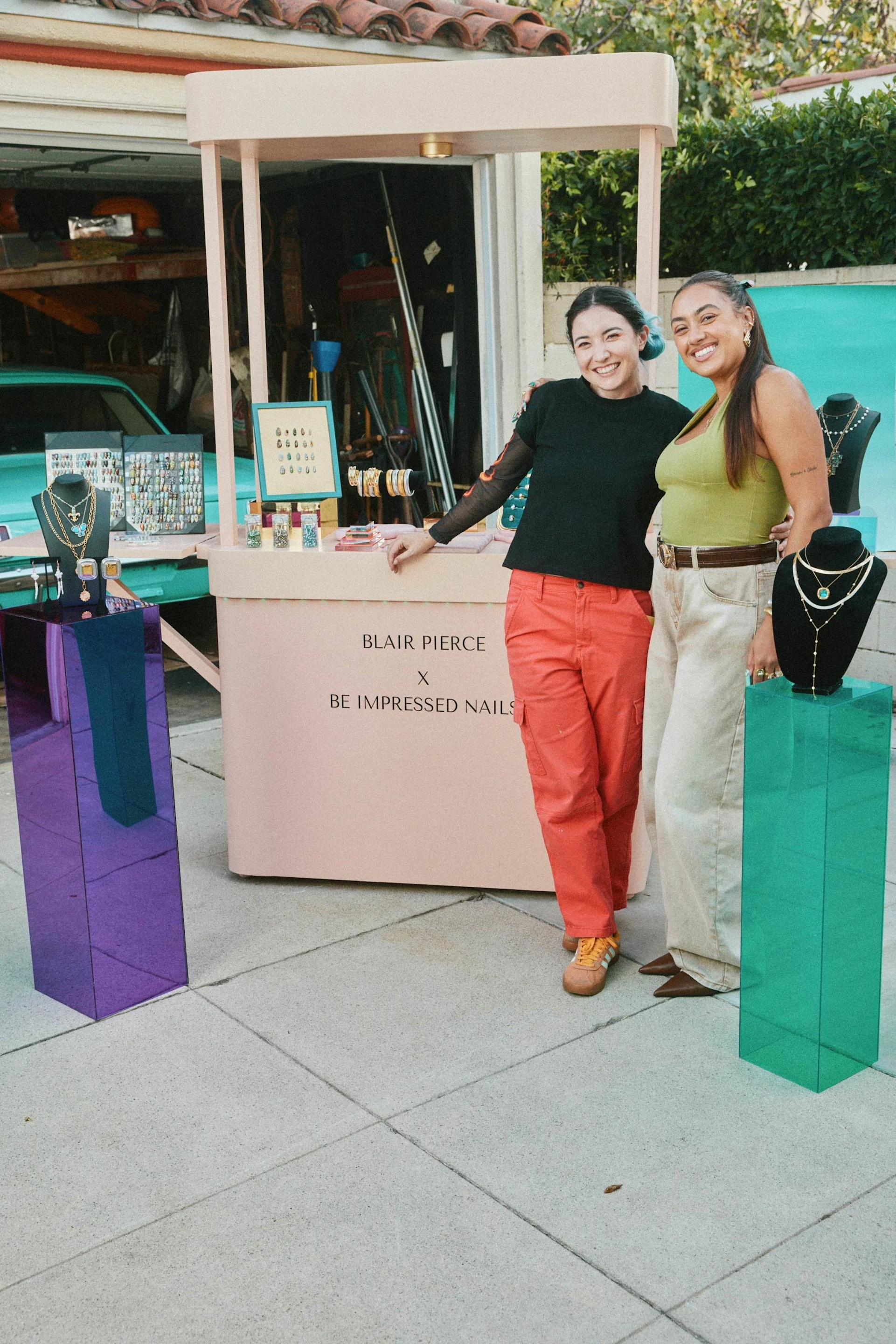 Two women smile and pose beside a small outdoor jewellery pop-up stall, with necklaces and earrings displayed on colourful stands.