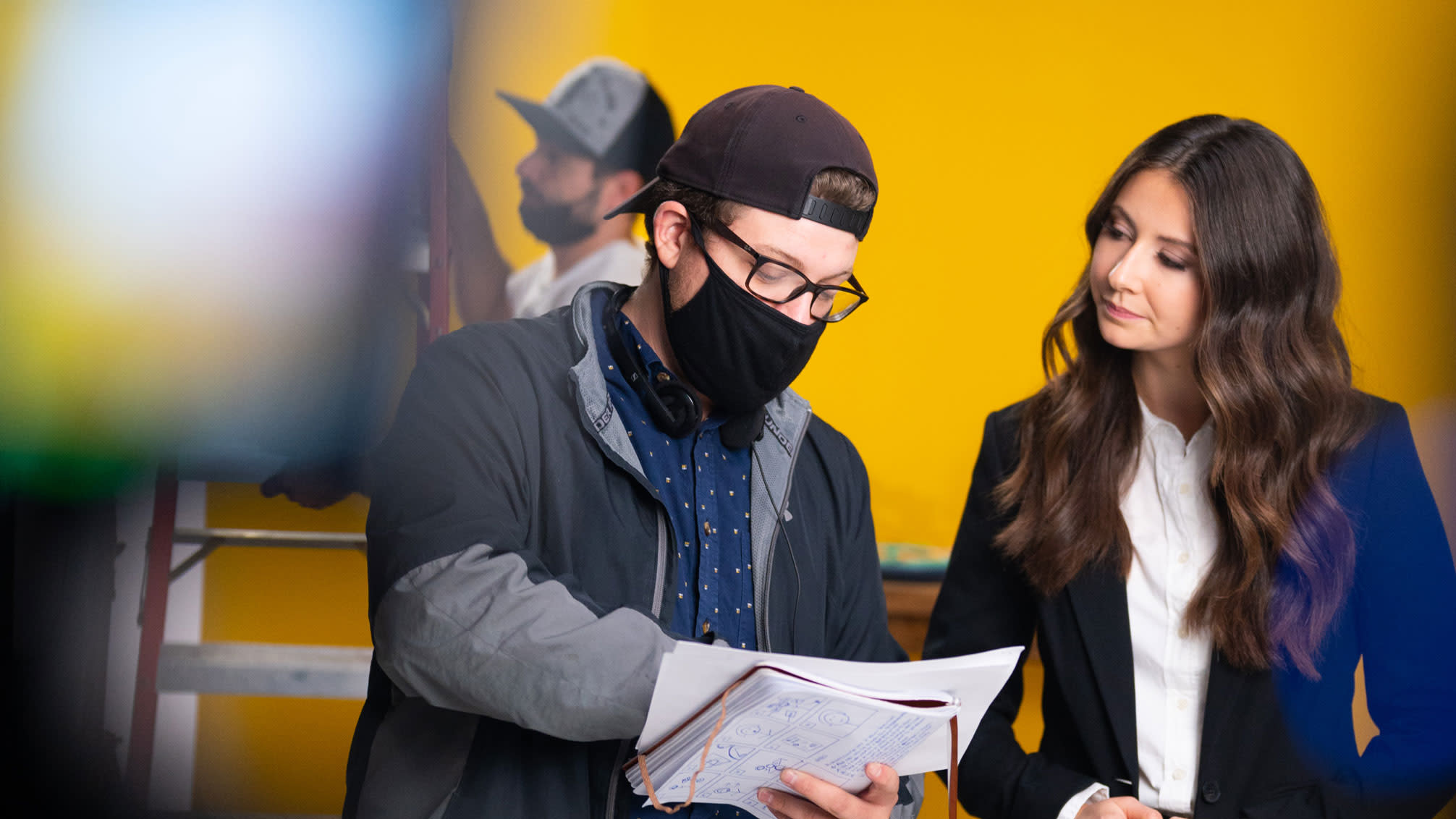 Oak + Rumble director, Brandon Hardy, on set during COVID, wearing a face mask, directing a female actress as they review the video script together.