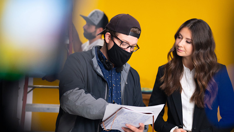 Oak + Rumble director, Brandon Hardy, on set during COVID, wearing a face mask, directing a female actress as they review the video script together.