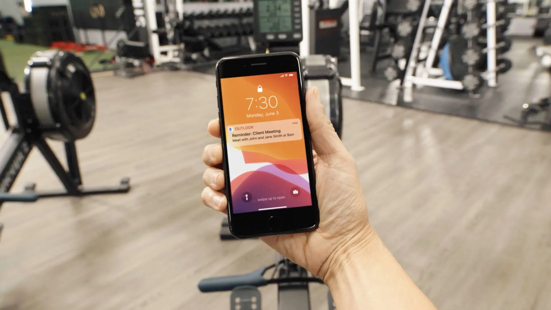 First-person view of a hand holding a phone showing a client meeting reminder, beside a rowing machine in a gym.