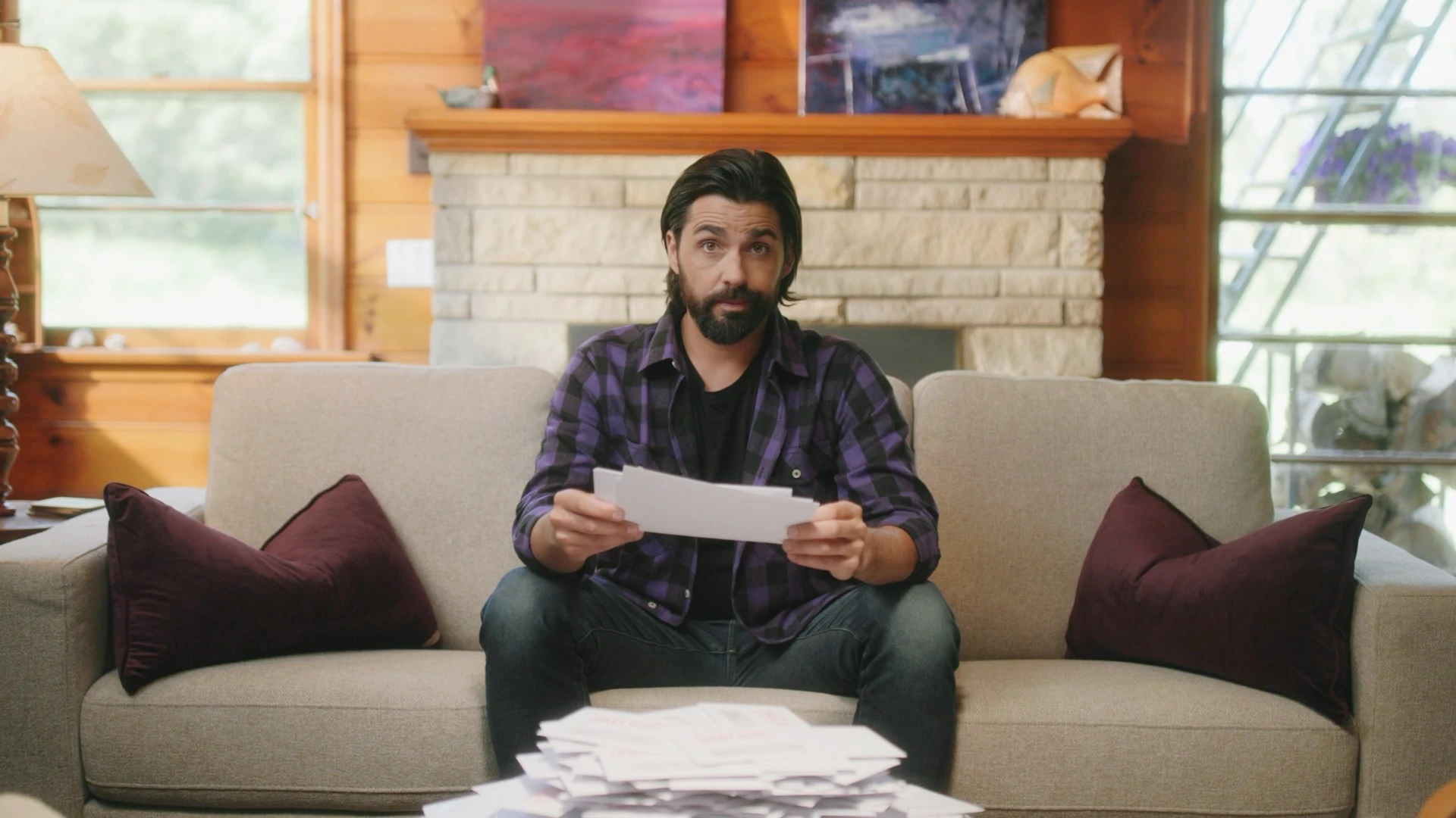 Actor in a purple plaid shirt on a cabin couch behind a pile of bills in Fairstone's Funny Canadian 2 commercial