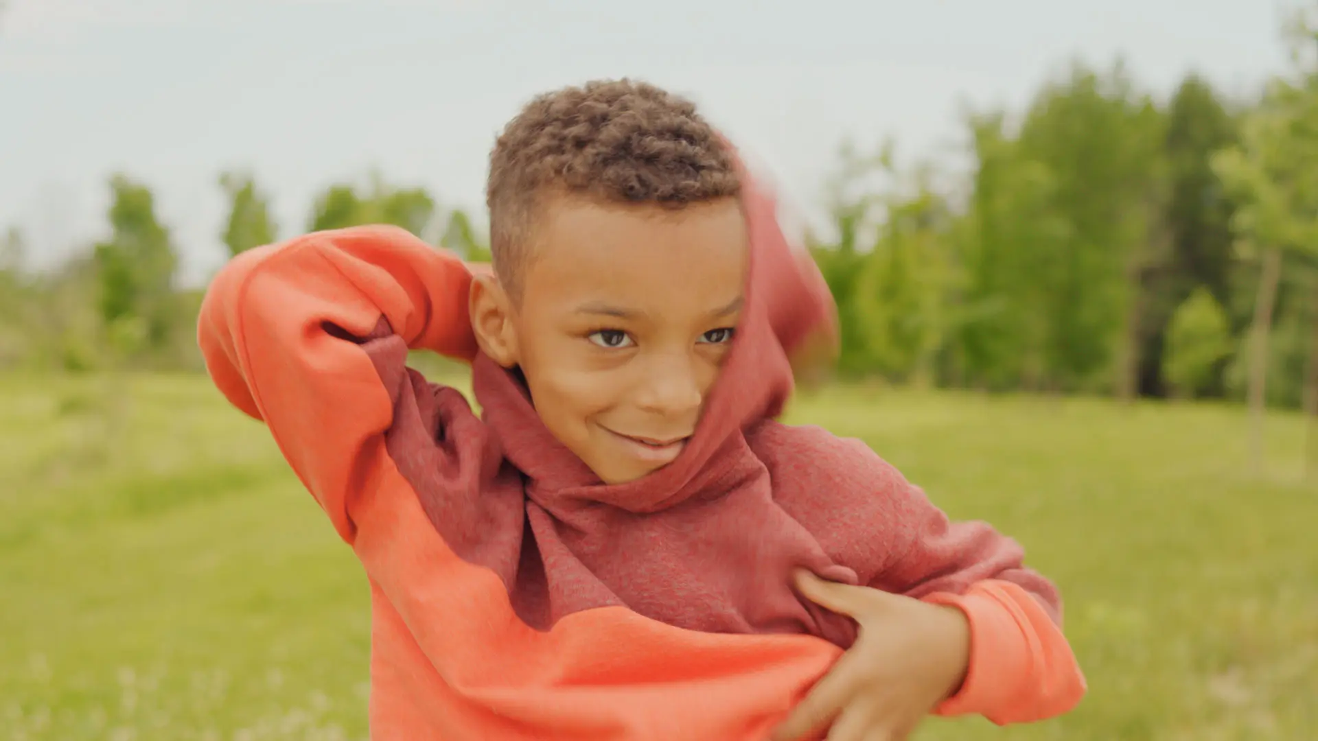 A smiling boy pulls an orange and red sweatshirt over his head in a park in an Oliver’s Labels commercial.