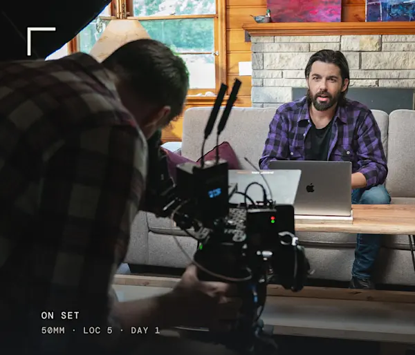 Behind the scenes with a camera operator in the foreground. Crew rigging a coffee table on plexiglass with strings to slide it into frame for Fairstone's Funny Canadian 2.