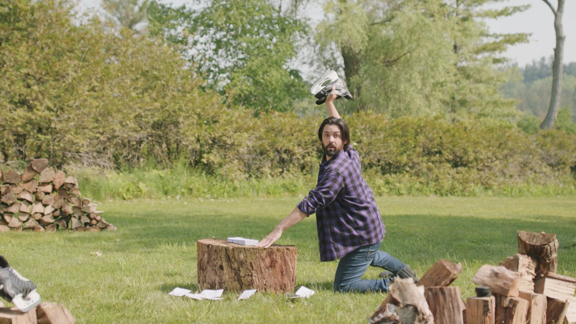 Gord is kneeling next to a tree stump, winding up to chop some bills up with a hockey skate in his hand.