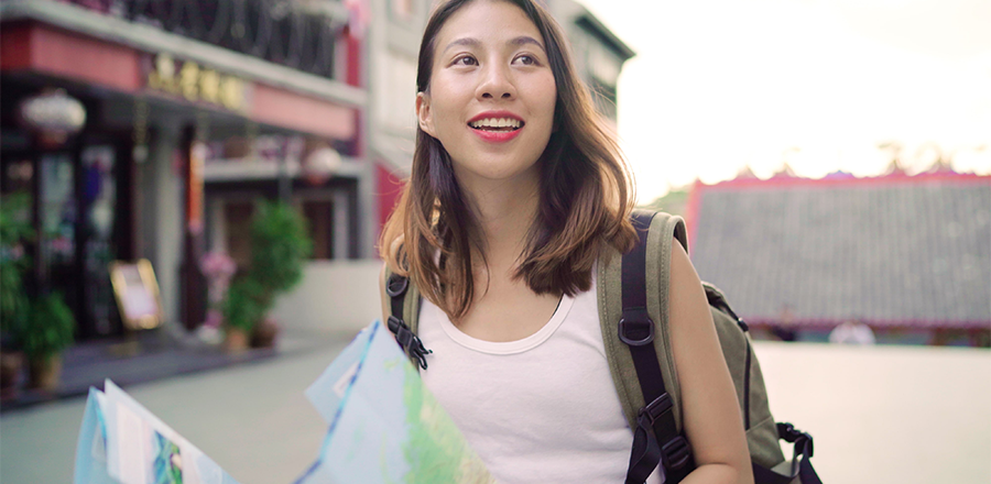 Una chica sonriente con una mochila sostiene un mapa desplegado. Al fondo se ven edificios y plantas en macetas.