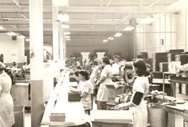 Fotografía en blanco y negro de una nave industrial. Mujeres con delantales blancos trabajan en la cinta transportadora.