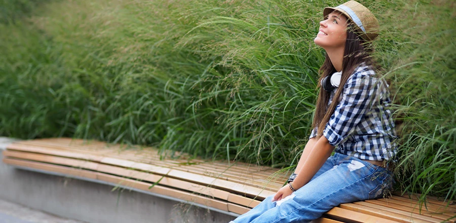 En un banco entre la vegetación una joven sentada con sombrero y auriculares en el cuello. Levanta la vista y sonríe.