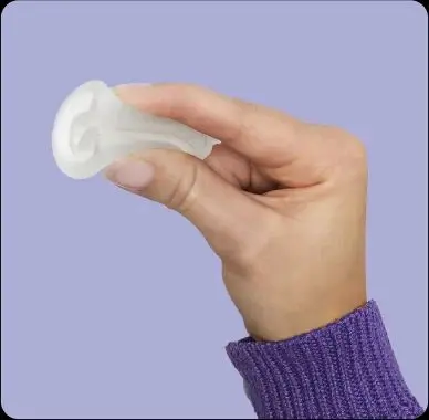 Sobre un fondo morado pálido, la mano de una mujer sostiene entre sus dedos una copa menstrual doblada y transparente.