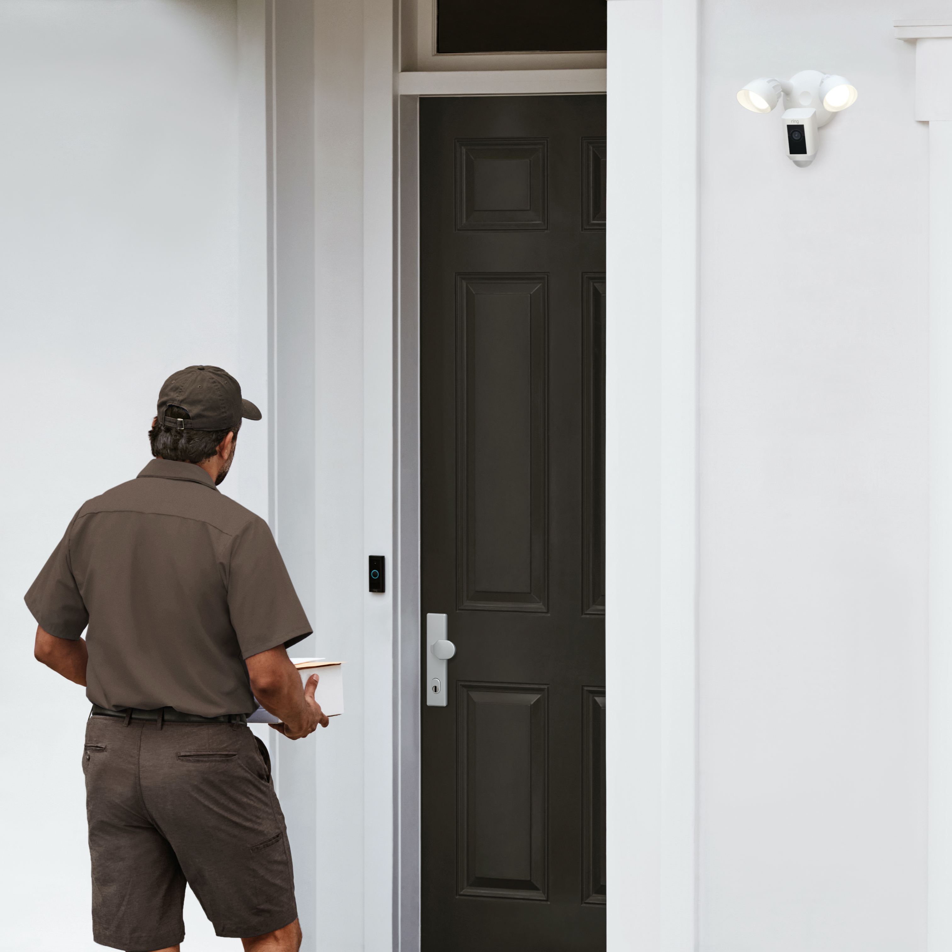 Delivery driver approaches door with parcel as white Ring Floodlight Cam monitors the entryway.