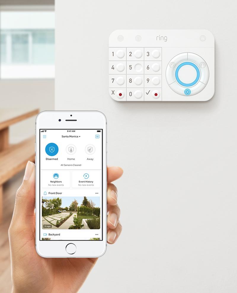 Person holds phone showing Ring app interface near mounted Ring Alarm Keypad on indoor wall.