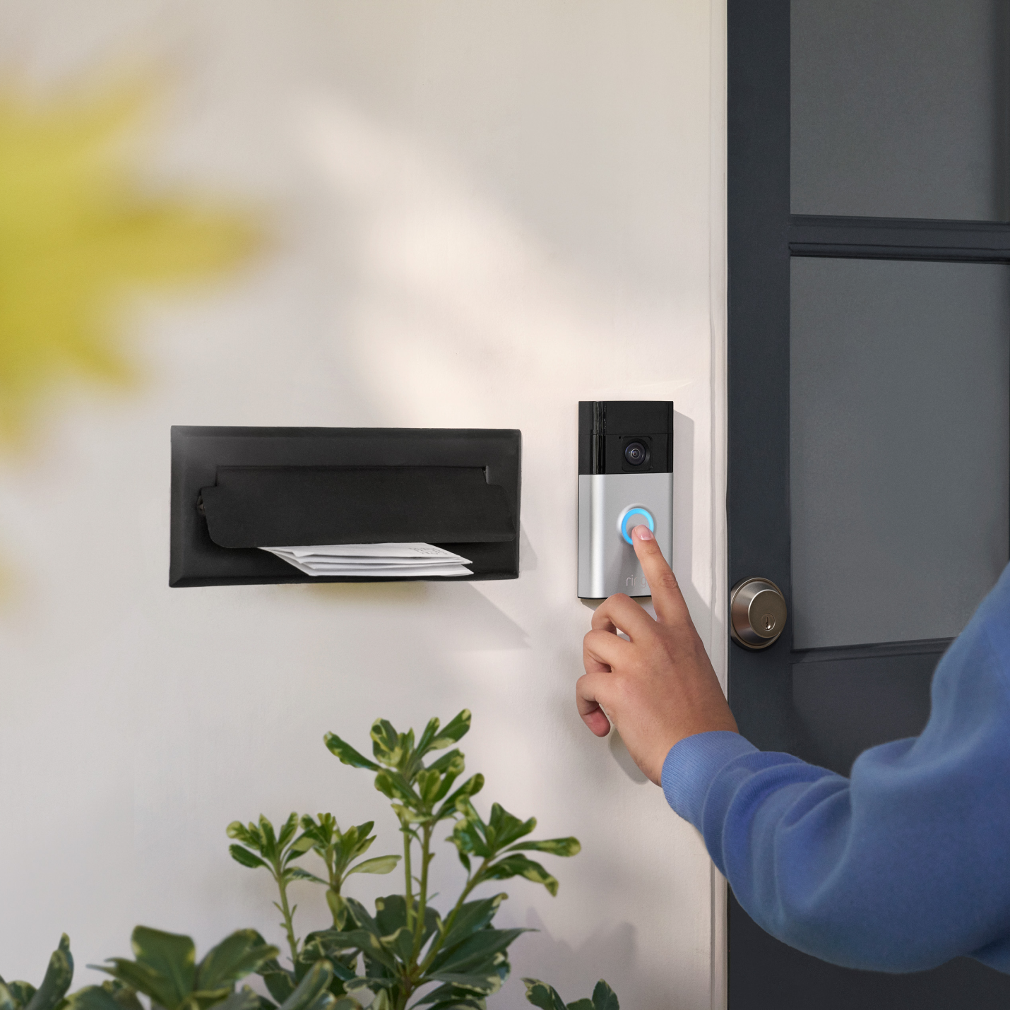 Person pressing illuminated Ring Video Doorbell button next to a front door and letterbox with visible mail.