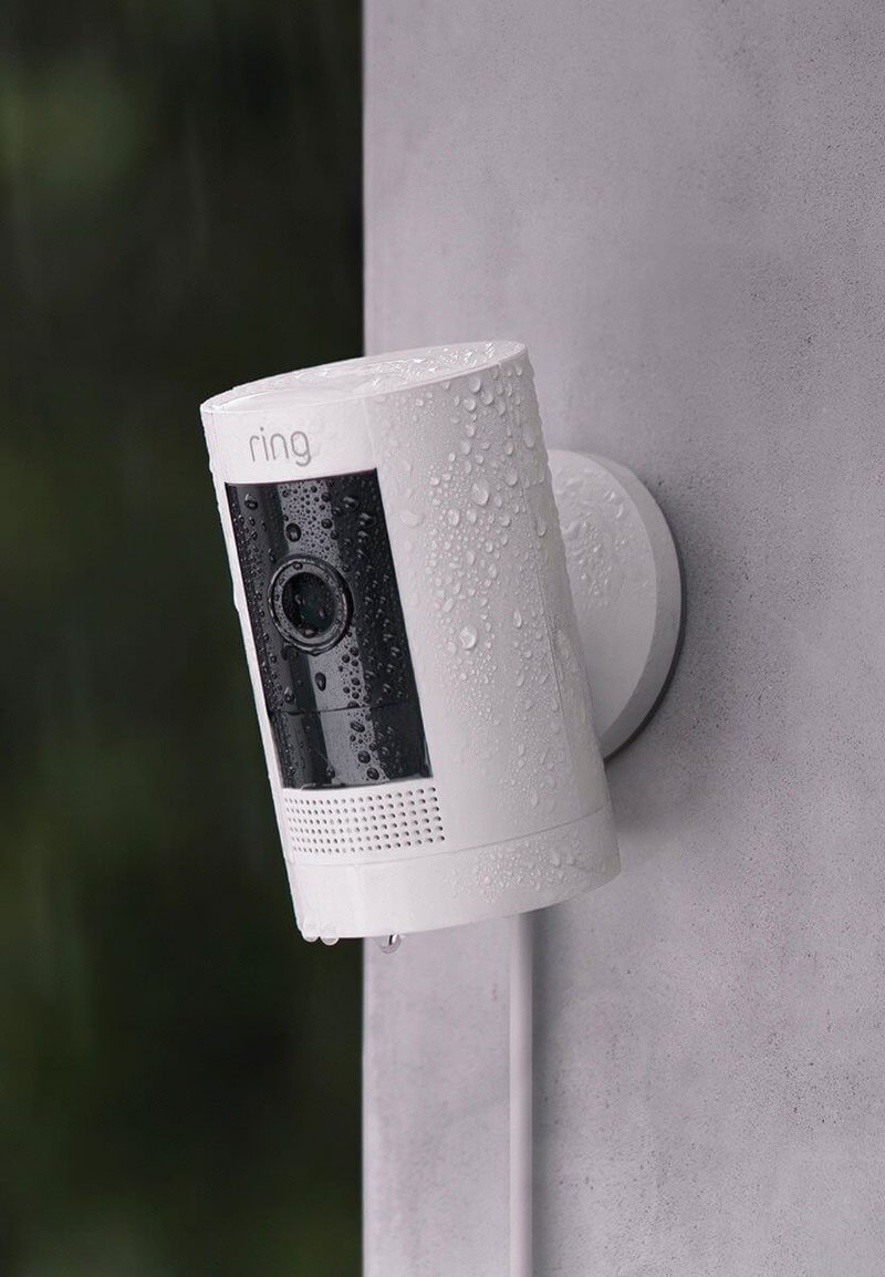 Weather-resistant Ring Stick Up Cam in white mounted outdoors with rain droplets on camera body and lens.