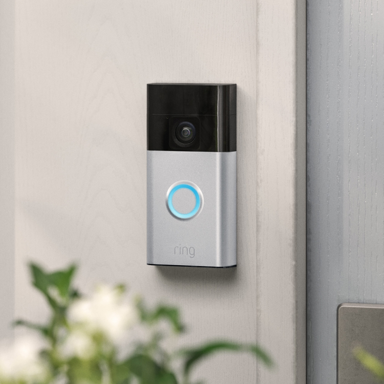 Ring Video Doorbell with illuminated button mounted beside a front door, surrounded by greenery and flowers.