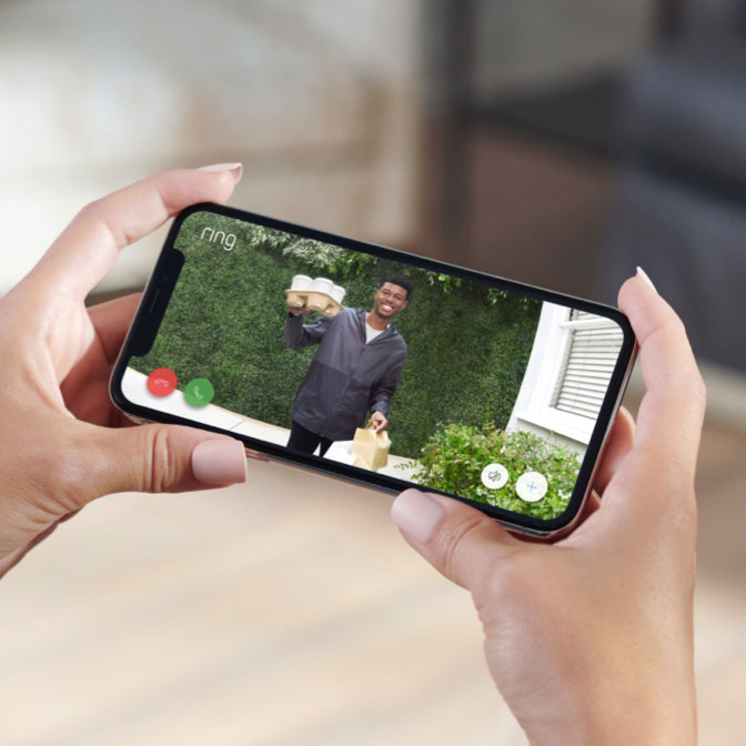 Person views live Ring doorbell video on smartphone screen of smiling delivery driver on porch holding food and drinks.