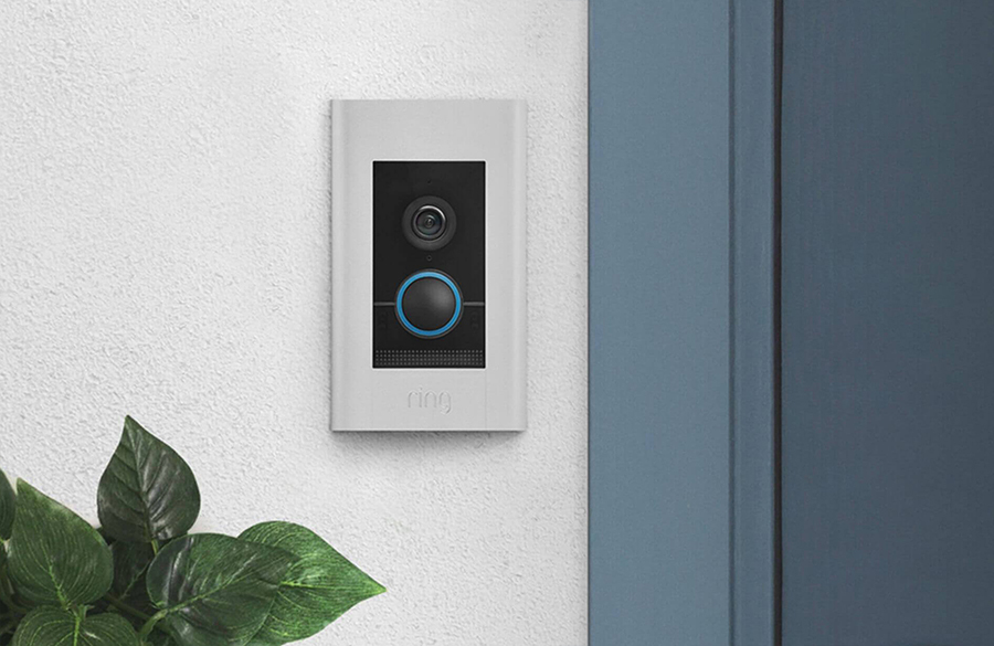 Ring Video Doorbell Elite mounted on a white textured wall next to a blue door, with plant leaves partially visible.