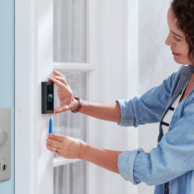 Woman installs Ring Video Doorbell Wired beside front door using screwdriver for secure mounting.