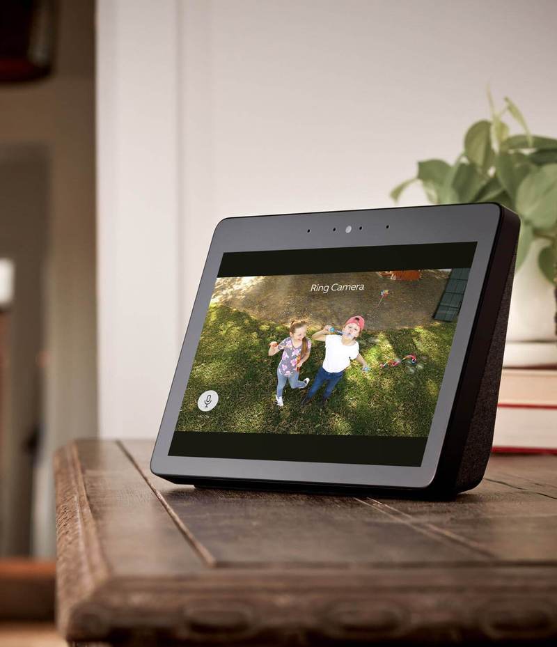 Echo Show on table displays Ring camera feed of two kids in yard, supporting hands-free monitoring with Alexa commands.