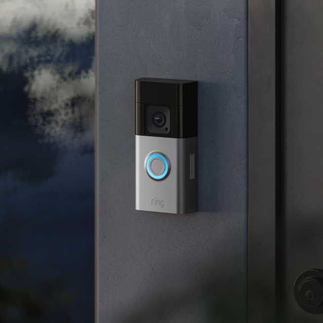 Ring Video Doorbell mounted on a dark exterior wall at dusk, with blue-lit button indicating power and readiness.