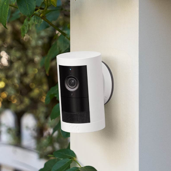 White Ring Stick Up Cam mounted on an exterior wall, positioned near green foliage for flexible outdoor security.