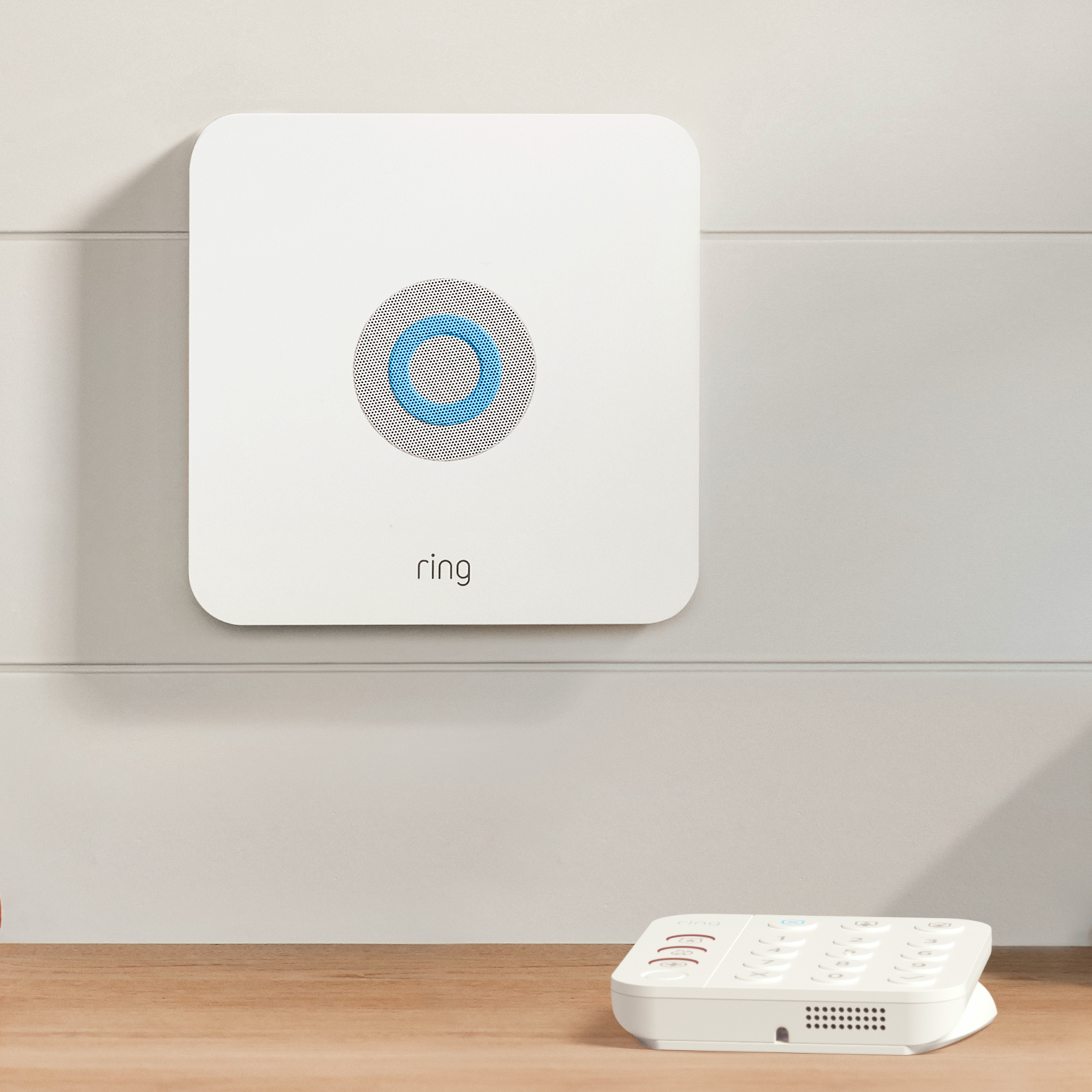 Ring Alarm base Station mounted on wall with illuminated blue ring, with Ring keypad on wooden surface below.