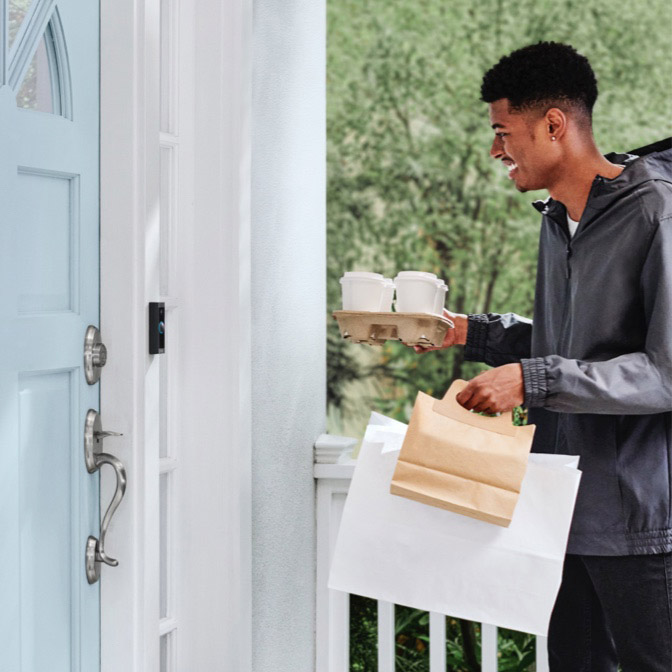 Delivery person approaches front door with food and drinks, seen by Ring Video Doorbell Wired on white door frame.
