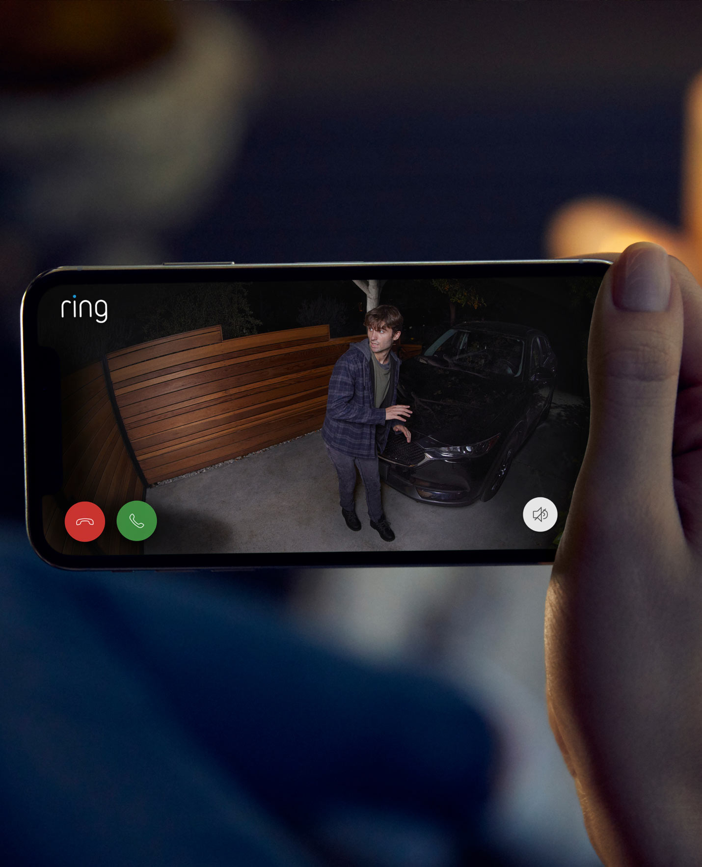 Live Ring app view of person near a parked car at night, spotlight on, with call and siren controls visible on screen.