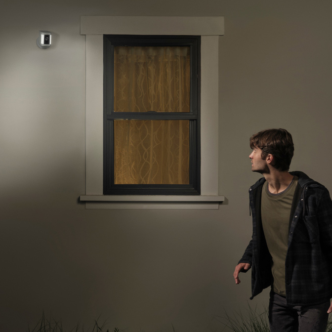 Man looks up at motion-triggered Spotlight Cam Plus as it illuminates him outside a house at night.