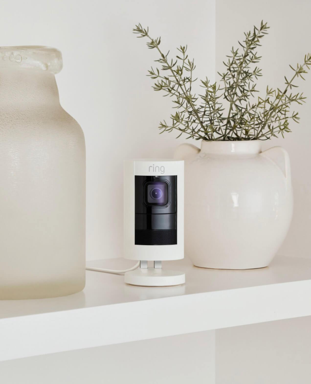 White Ring Stick Up Cam placed on white shelf between frosted glass jar and ceramic vase with greenery.